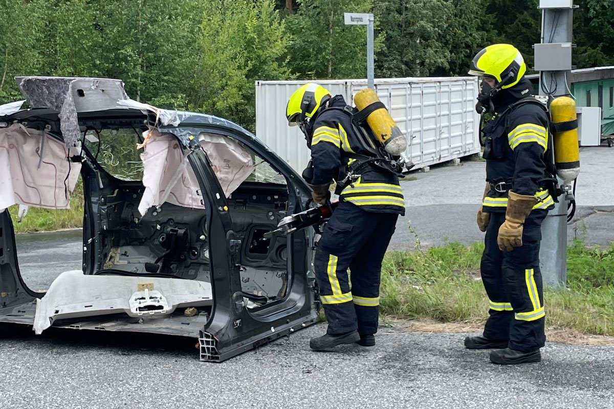 Kaksi henkilöä sammutusasuissa, toinen heistä leikkaa auton runkoa.