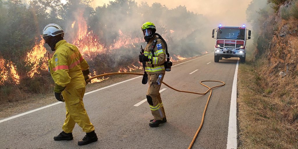 Kaksi pelastajaa ja sammutusauto tiellä, tien vieressä palaa tulipalo.