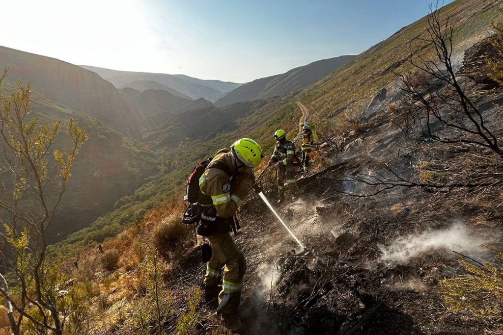 Kolme sammutusasuista henkilöä jälkisammuttamassa maastopaloa vuorenrinteessä, taustalla vuorimaisemaa.