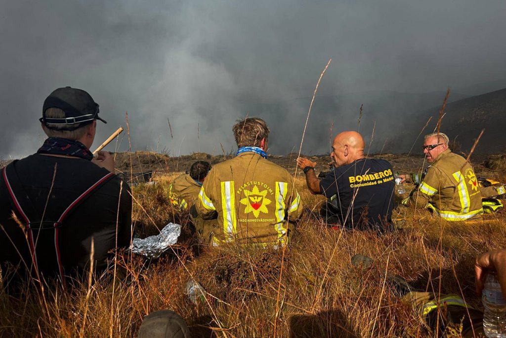 Viisi pelastusalan henkilöä istumassa ruohikolla ja keskustelemassa, taustalla savuinen vuorimaisema.