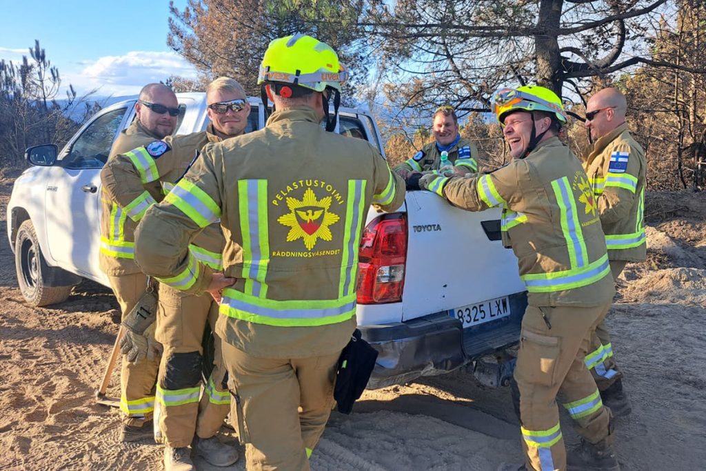 Kuusi sammutusasuista henkilöä keskustelemassa lava-auton ympärillä, taustalla vuorimaisemaa.