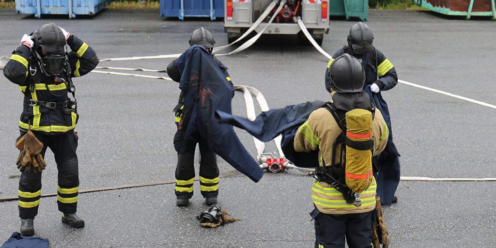 Pelastustoimen henkilöstöä sammutusasuissa, taustalla sammutusauto ja sammutusletkuja.