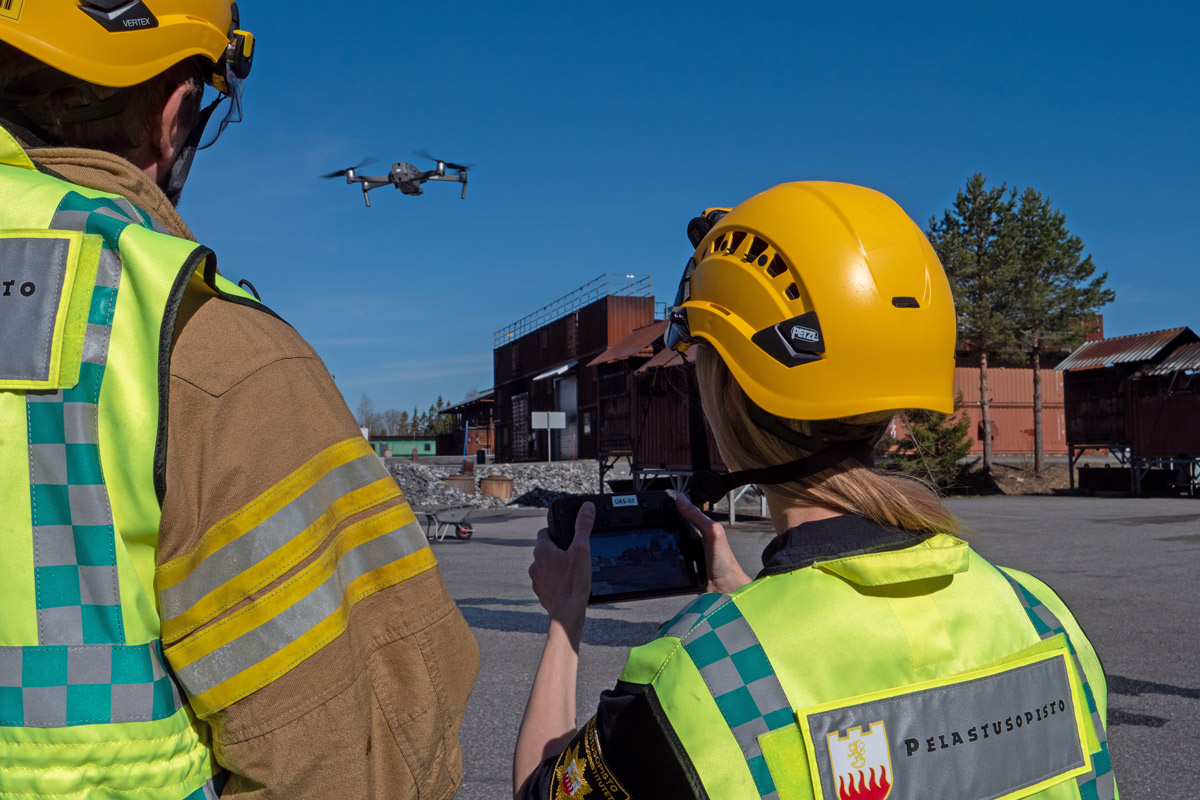 Kaksi henkilöä lennättää dronea, henkilöiden selät etualalla, drone taustalla ilmassa.