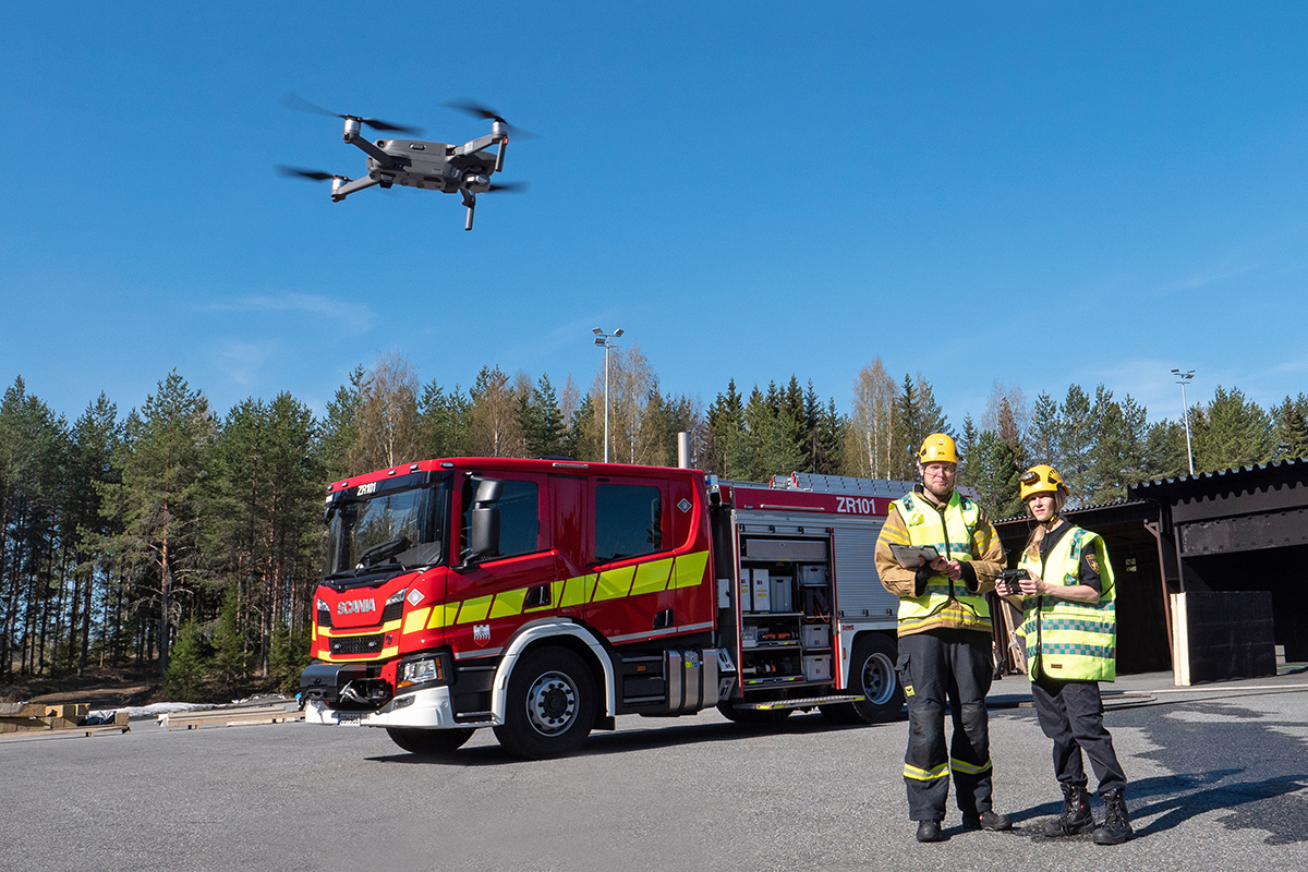 Kaksi henkilöä lennättää dronea, taustalla sammutusauto.