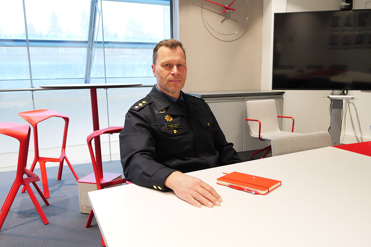 Virkapukuinen henkilö istuu neuvottelutilassa pöydän ääressä ja katsoo kameraan.