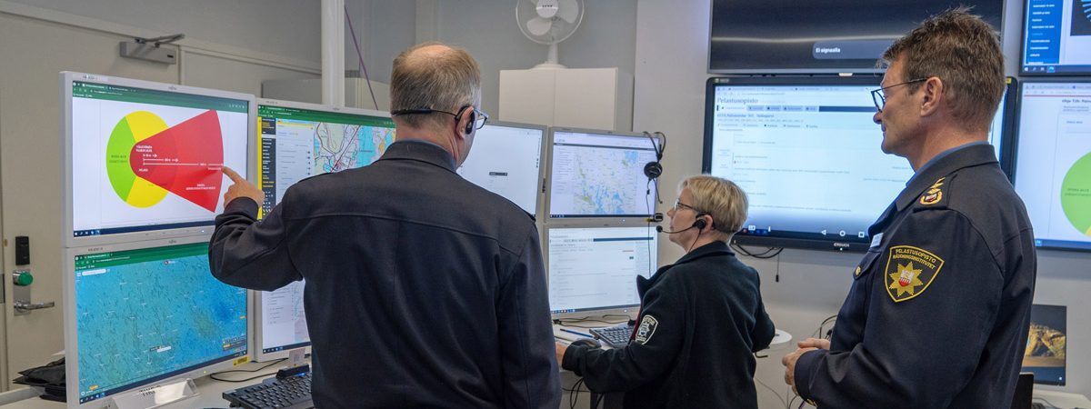 Kolme pelastusasuista henkilöä katselevat useaa tietokoneen näyttöä.
