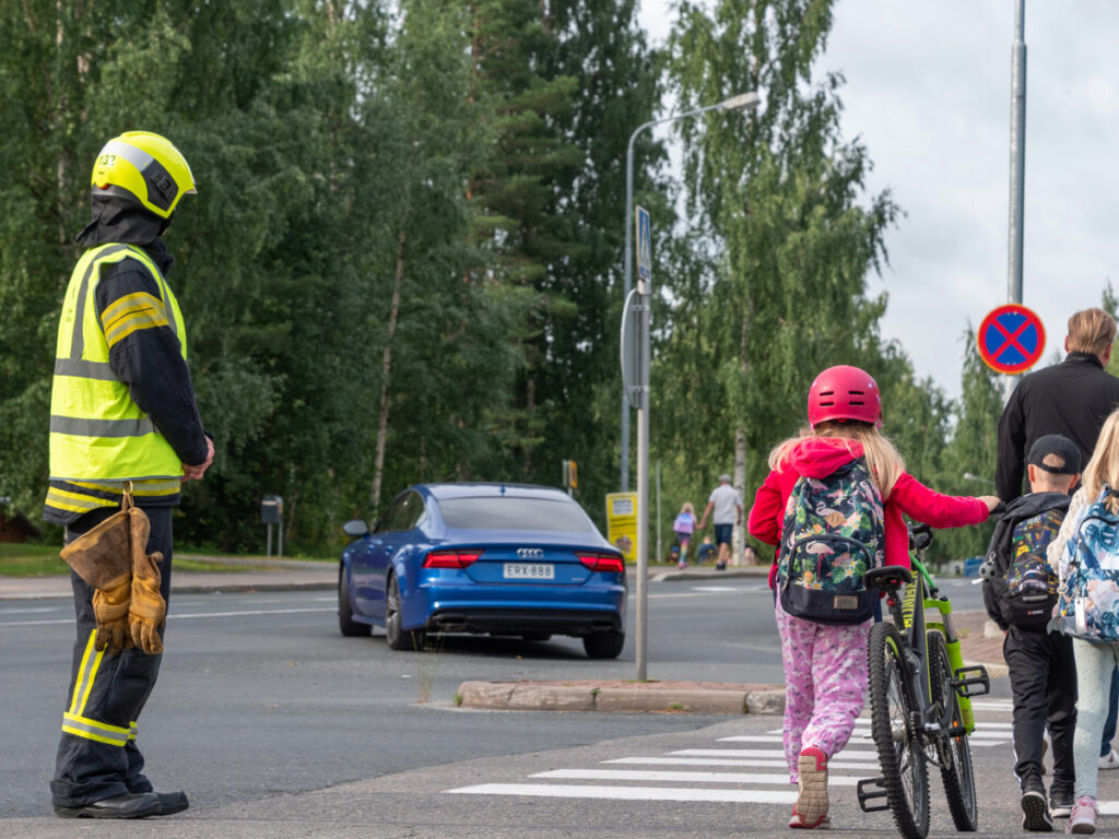 Pelastaja katsoo suojatien vieressä, kun koululaiset ylittävät pyörää taluttaen tietä.