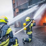 Kolme sammutusasuista henkilöä suihkuttamassa vesiletkulla vettä rakennukseen. Rakennuksen ovet on auki ja sisällä palaa tuli.