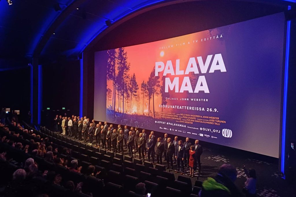 Yleisöä elokuvateatterin salissa, salin edessä henkilöitä rivissä valkokankaan edessä. Kankaalla näkyy elokuvan tiedot.