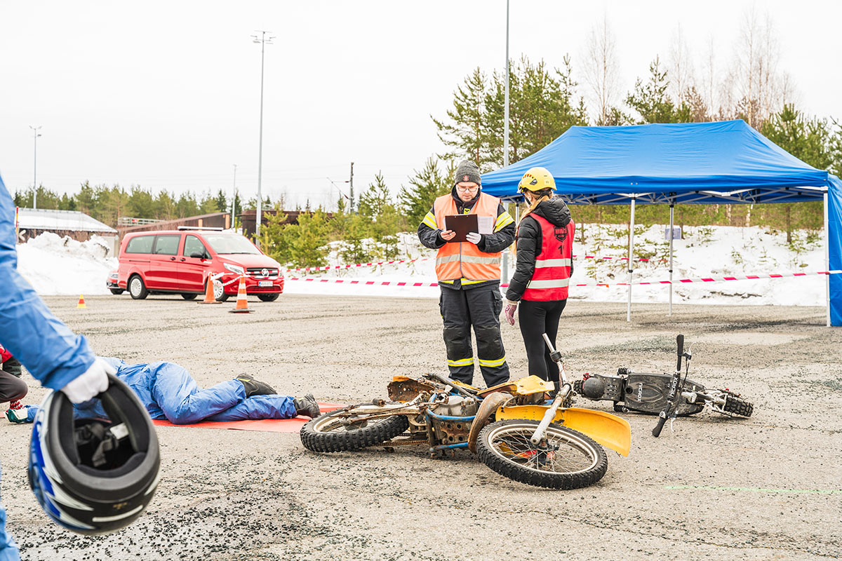Henkilö keskustelee Nouhätä!-rastin ohjaajan kanssa, maassa mopo kyljellään ja henkilö vieressä kylkiasennossa.