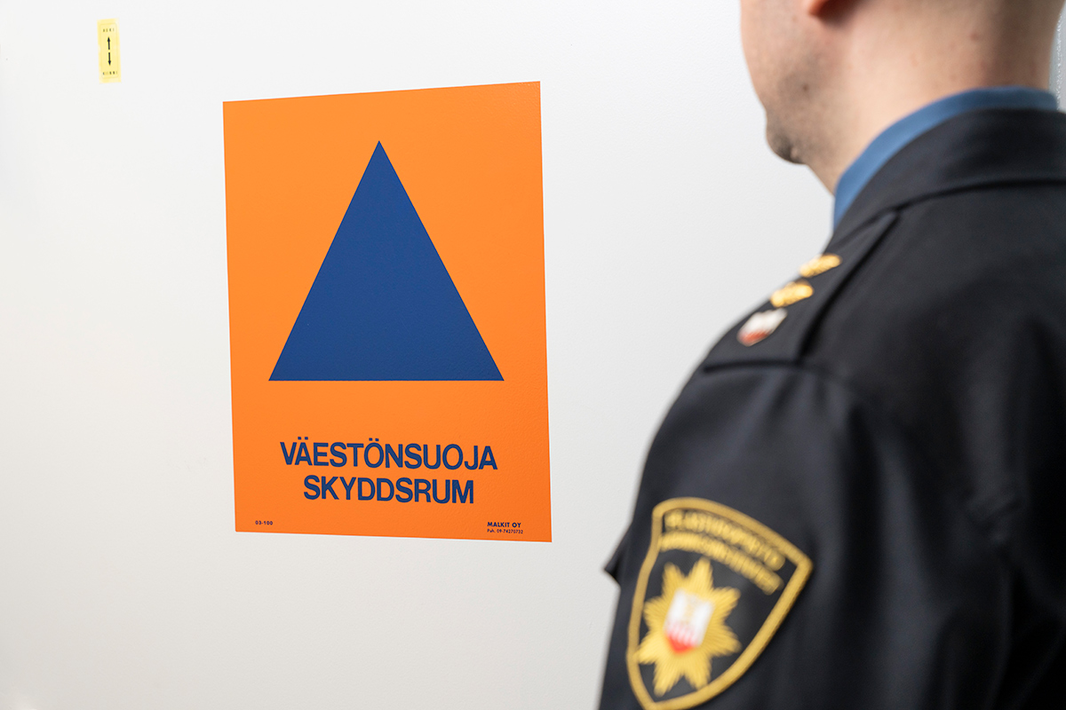 Virkapukuinen henkilö väestönsuojan oven edessä, ovessa oranssi-sininen väestönsuojan logo.