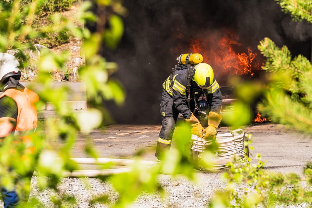 Sammutusasuinen henkilö avaamassa sammutusletkua säilytyskehikosta. Taustalla henkilöauto, joka on syttynyt tuleen.