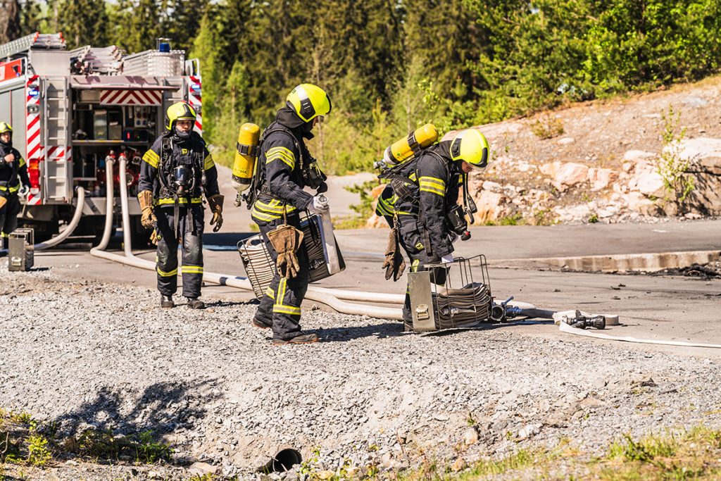 Neljä pelastajaopiskelijaa sammutusasuissa kantamassa letkukehikoita, taustalla sammutusauto.