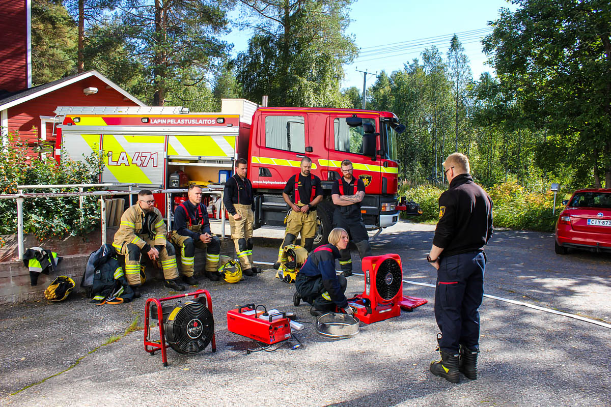 Taustalla sammutusauto, jonka edessä sopimushenkilöstöryhmä ja työvälineitä. Ryhmän edessä ohjaaja.