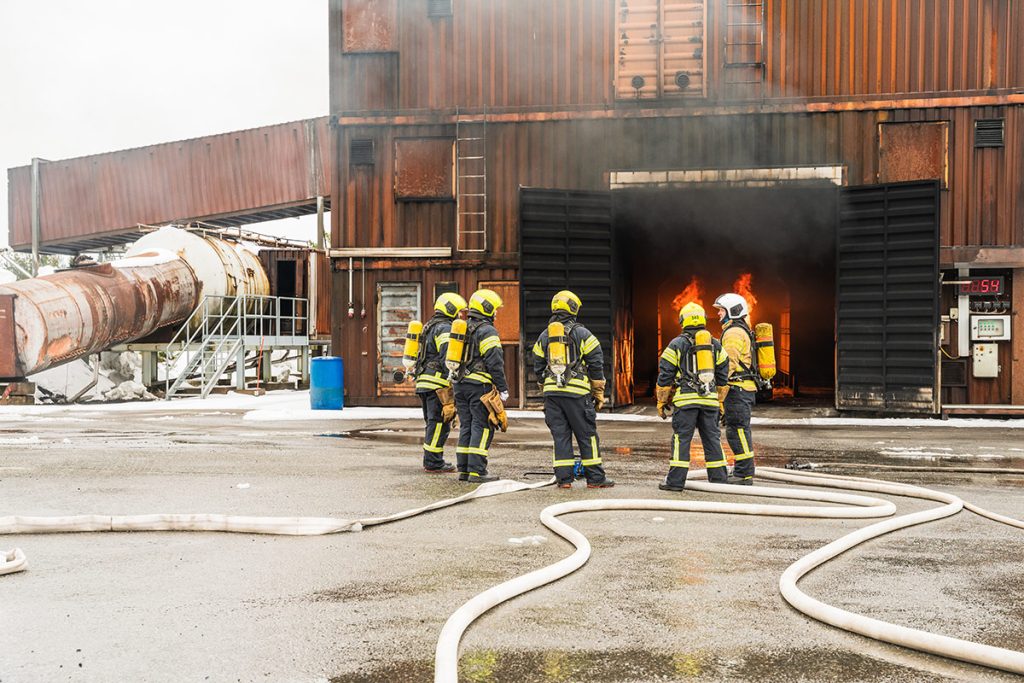 Pelastushenkilöstöä Sammutustekniikkakurssin vastaavan kouluttajan koulutuksesa Pelastusopiston harjoitusalueella. Kurssilaisia kokoontuneena ohjaajan luo, taustalla rakennus, jonka avoimista ovista näkyy tulipalo.