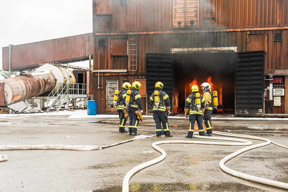 Pelastushenkilöstöä Sammutustekniikkakurssin vastaavan kouluttajan koulutuksesa Pelastusopiston harjoitusalueella. Kurssilaisia kokoontuneena ohjaajan luo, taustalla rakennus, jonka avoimista ovista näkyy tulipalo.