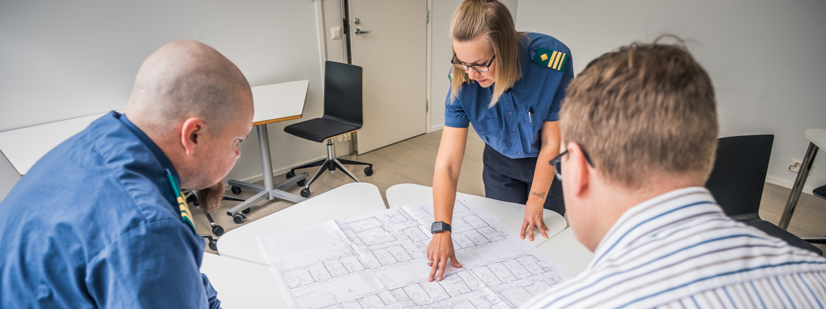 Kaksi virkapukuista ja yksi siviiliasuinen henkilö tarkastelevat suurta karttaa.