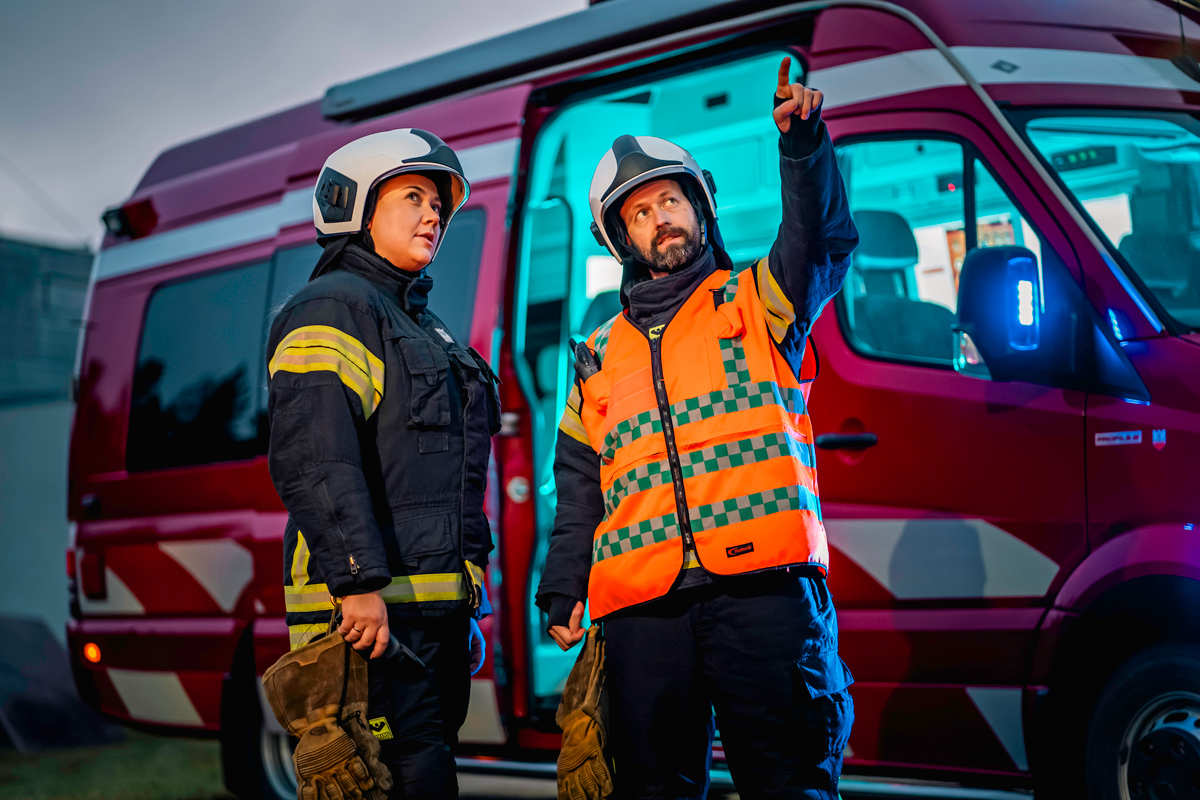 Kaksi henkilöä päällystöstä seisoo auton edessä, he katsovat samaan suuntaan oikealle ylöspäin.