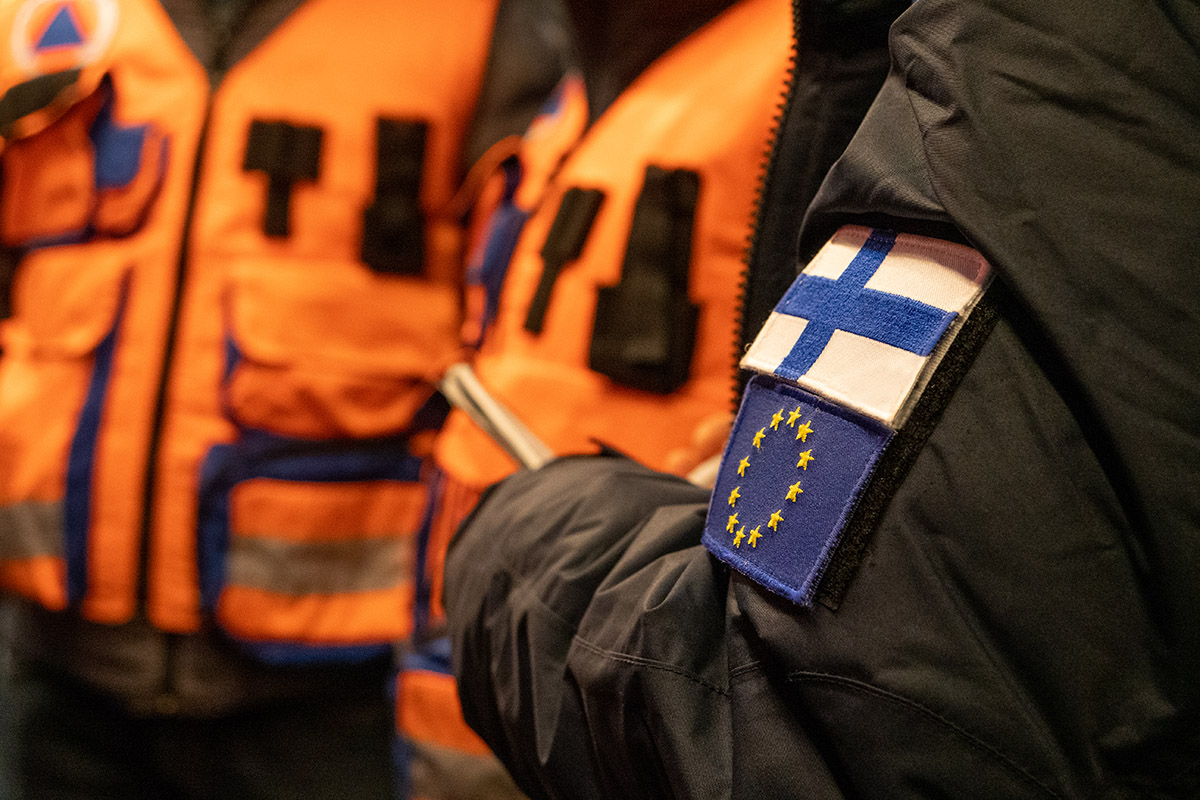 Kansainvälisen pelastustoiminnan henkilöstöä rivissä. Lähikuvassa takin hiha, jossa suomen ja Euroopan unionin liput.