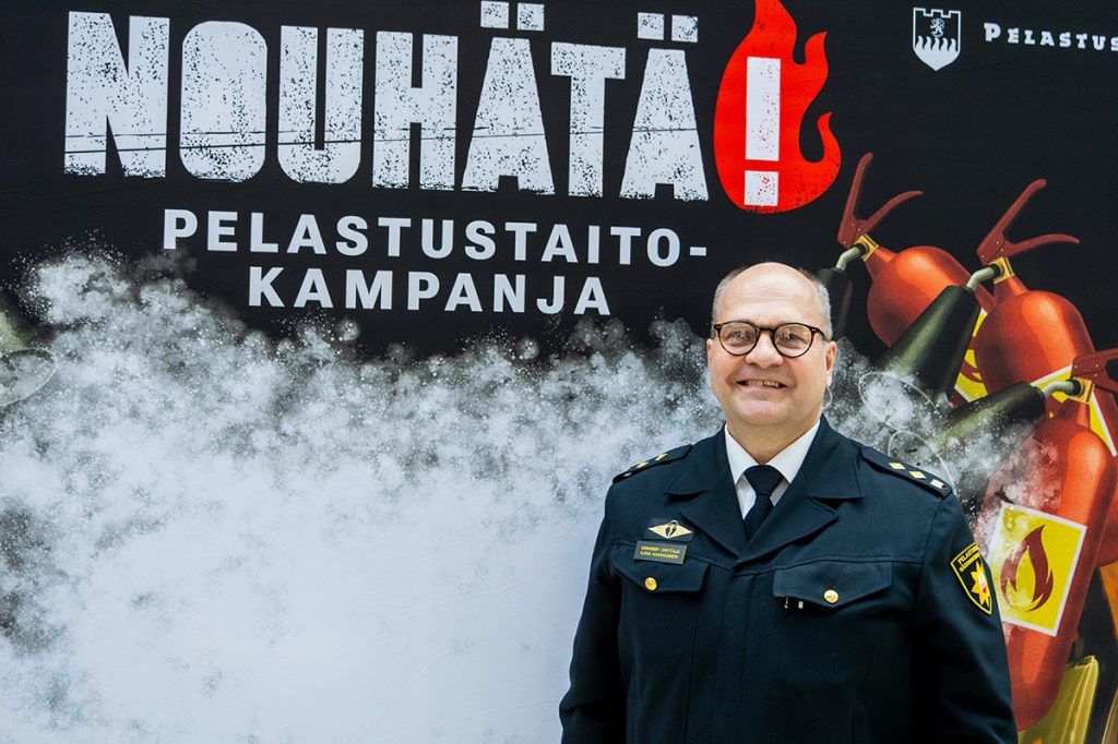Virka-asuinen henkilö hymyilee ja katsoo kameraan, taustalla NouHätä!-pelastustaitokampanjan kuvaseinä.