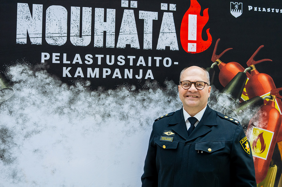 Virka-asuinen henkilö hymyilee ja katsoo kameraan, taustalla NouHätä!-pelastustaitokampanjan kuvaseinä.