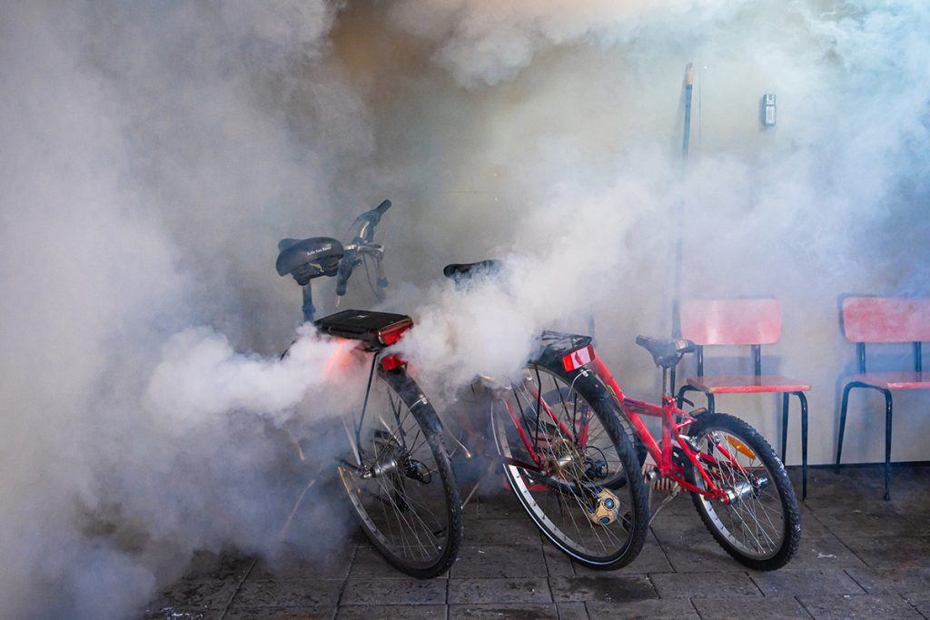 Sähköpyöriä pysäköitynä rakennuksen seinustalle. Yhden pyörän akku on syttynyt palamaan, tilassa on paljon savua.