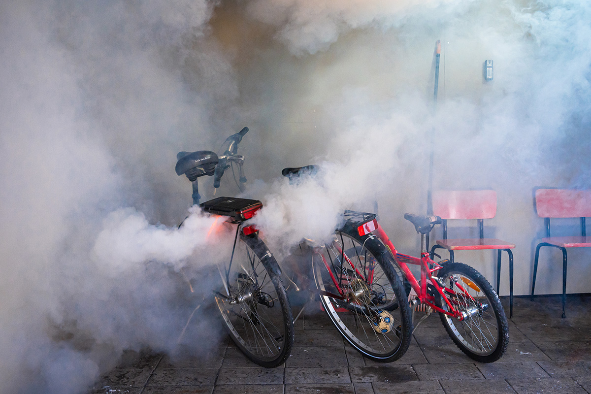 Sähköpyöriä pysäköitynä rakennuksen seinustalle. Yhden pyörän akku on syttynyt palamaan, tilassa on paljon savua.
