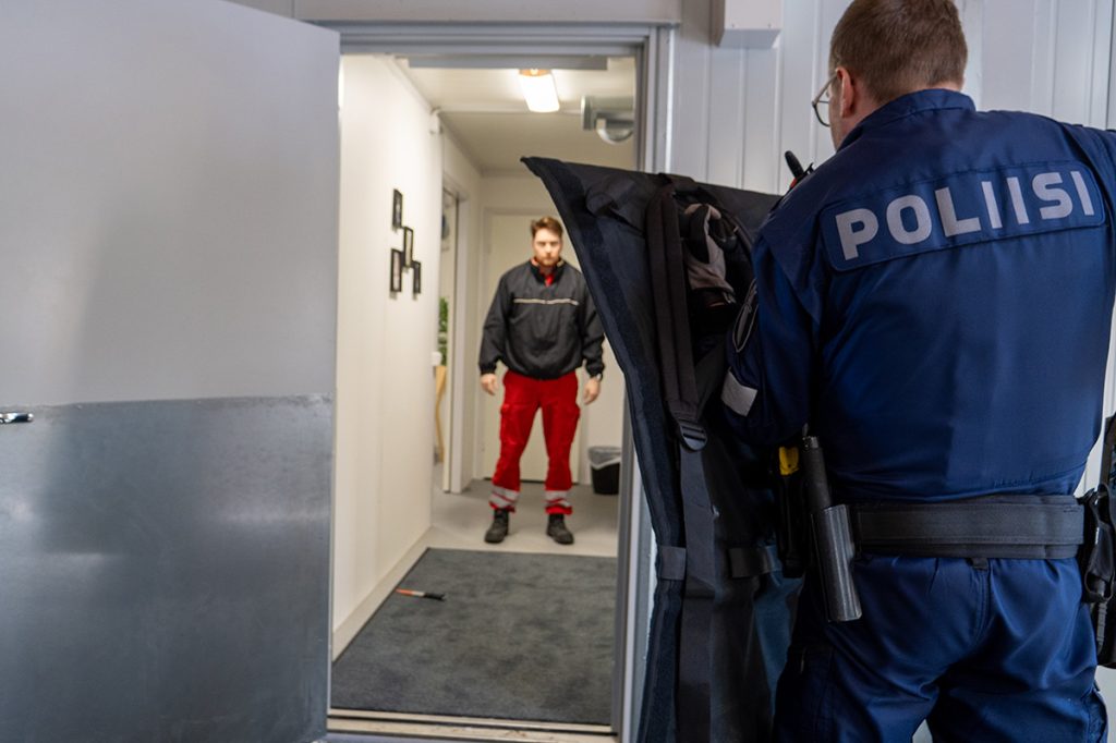 Etualalla poliisi seisoo rakennuksessa oven suulla ja suojaa itseään, taustalla näkyy käytävä ja käytävällä seisoo henkilö, joka katsoo poliisiin päin.