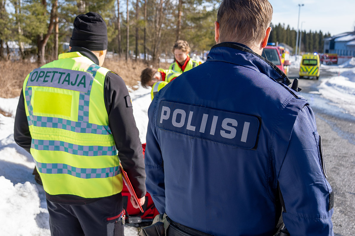 Opettaja ja poliisi vierekkäin ulkona seuraamassa ensihoidon harjoitustilannetta, henkilöt ovat selkä kuvaan päin.