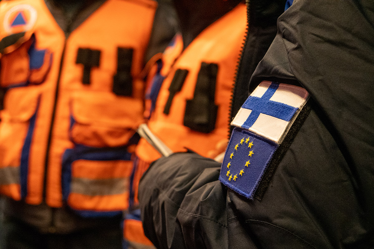 Takin hiha, jossa Suomen lippu ja Euroopan unionin lippu. Taustalla kaksi oranssitakkista henkilöä.