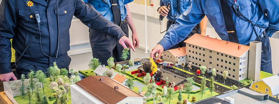 Lähikuva pelastusasuisen henkilöiden käsistä pienoismallin ympärillä, jossa rakennuksia, maastoa ja paloautoja.