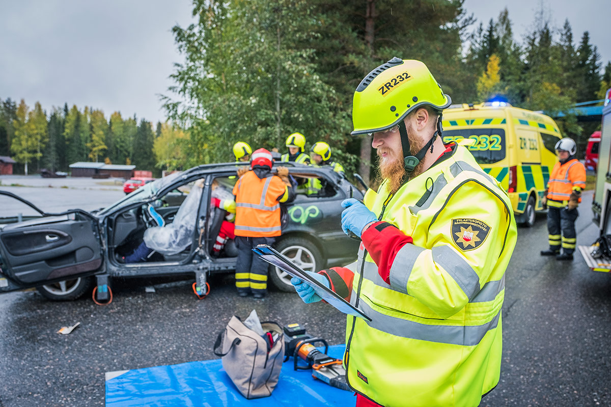 Henkilö puhuu Virve-puhelimeen, taustalla pelastushenkilöstöä pelastamassa henkilöä kolariin joutuneesta henkilöautosta.