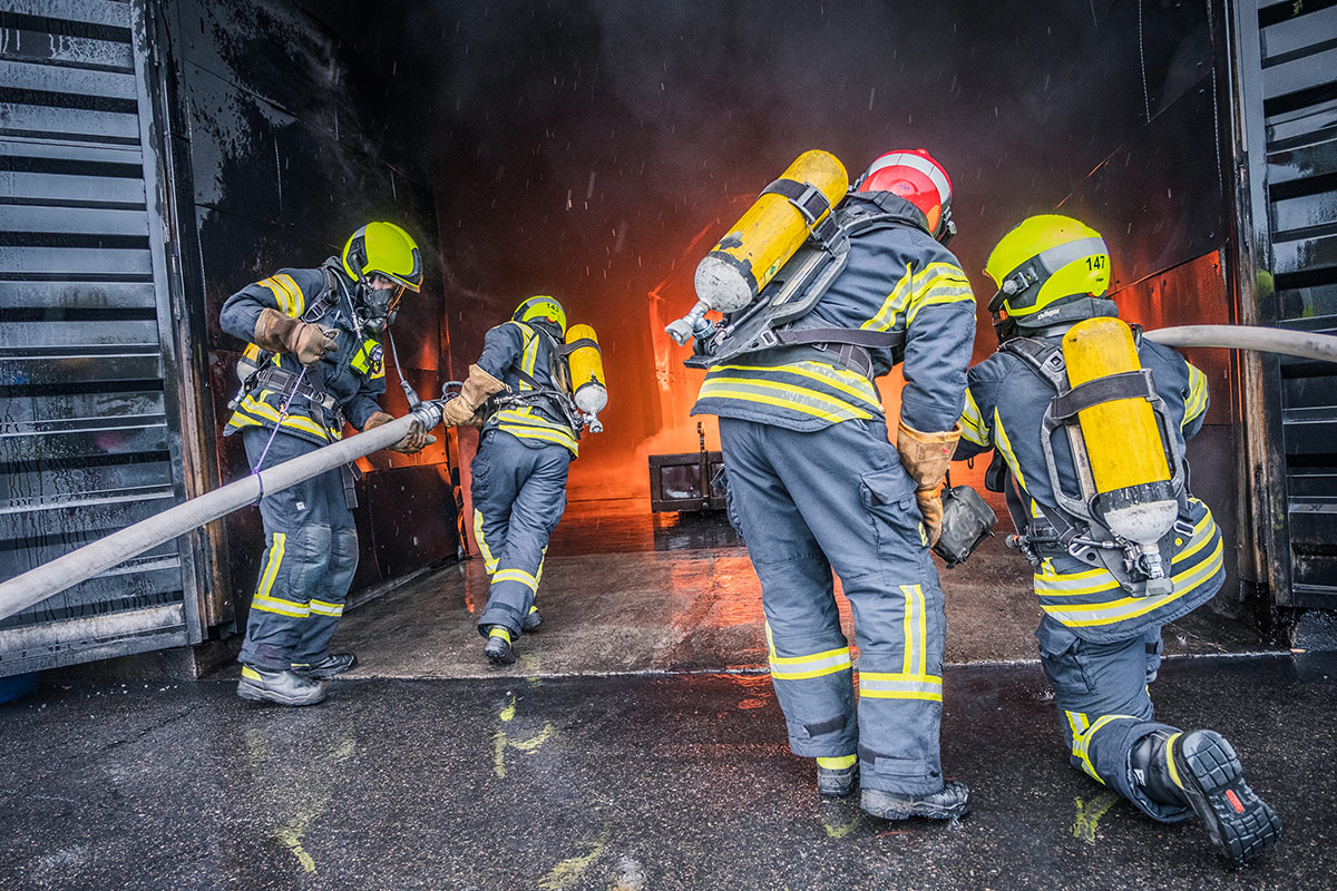 Kolme pelastajaa ja yksi alipäällystön edustaja sammutusletkujen kanssa rakennuksen edustalla. Rakennuksen ovi on auki ja sisällä palaa tulipalo.