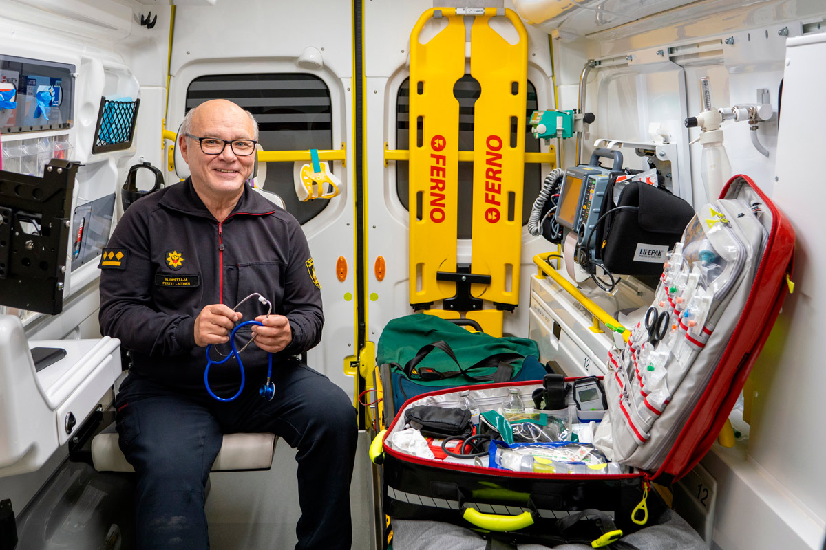 Pertti Laitinen istuu ambulanssissa esittelemässä ensiaputarvikkeita. Laitinen hymyilee ja katsoo kameraan.