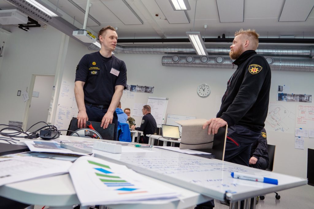 Henkilöitä Pelastusopiston luokkatilassa johtamisen pilottikoulutuksessa, etualalla kaksi henkilöä keskustelee pöydän äärellä.