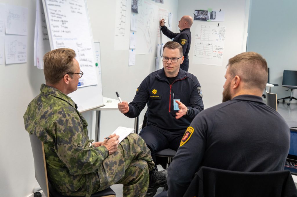 Henkilöitä Pelastusopiston luokkatilassa johtamisen pilottikoulutuksessa, etualalla kolme henkilöä keskustelee keskenään.