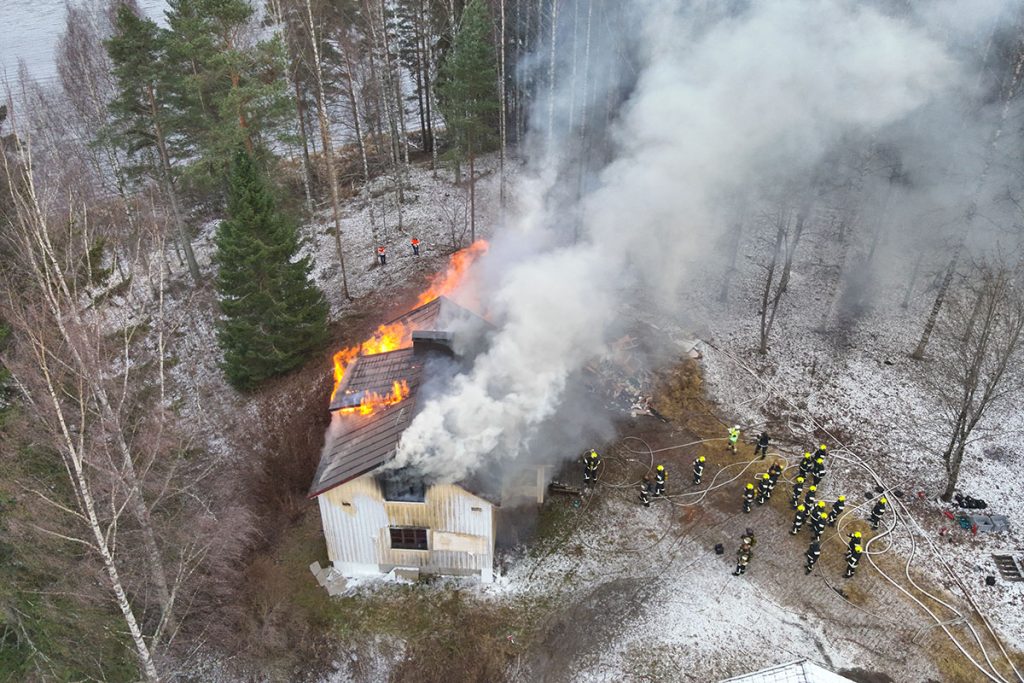 Ilmakuva palavasta talosta, pelastushenkilöstöä pihalla sammuttamassa paloa. Alueen ympärillä on metsää ja vesistöä.