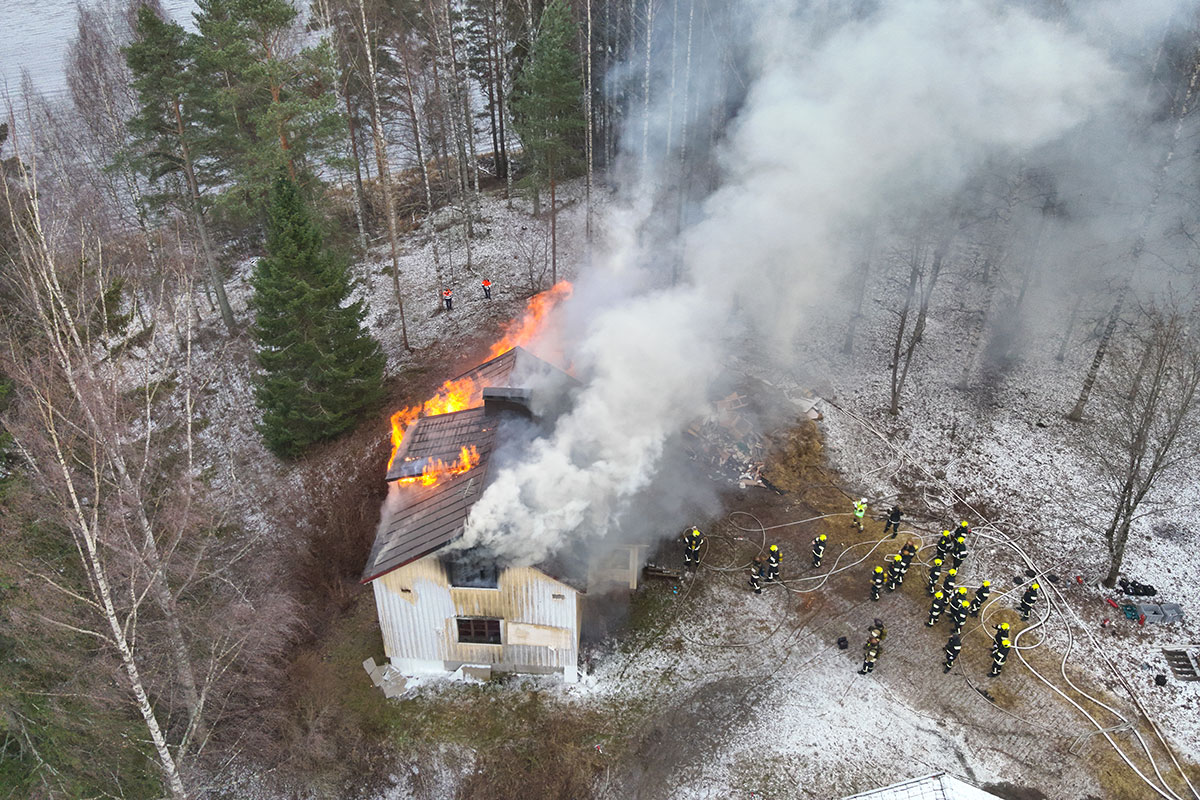 Ilmakuva palavasta talosta, pelastushenkilöstöä pihalla sammuttamassa paloa. Alueen ympärillä on metsää ja vesistöä.