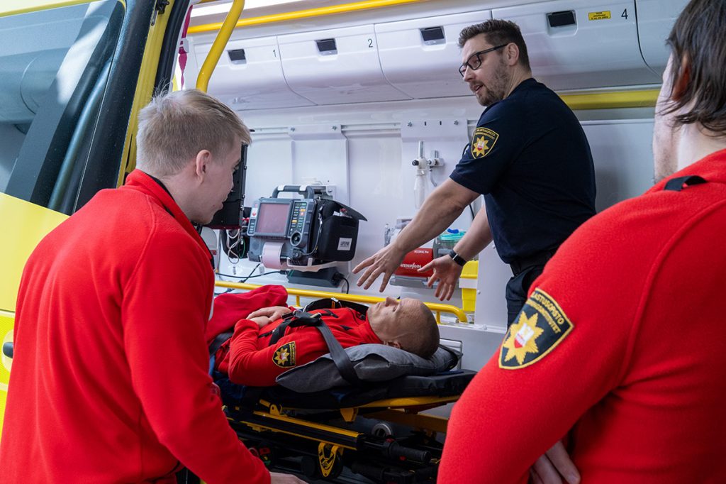 Opettaja ohjaa pelastajaopiskelijoita ambulanssissa. Yksi opiskelija makaa paareilla ja kaksi muuta opiskelijaa seuraa tilannetta ambulanssin oven suussa.