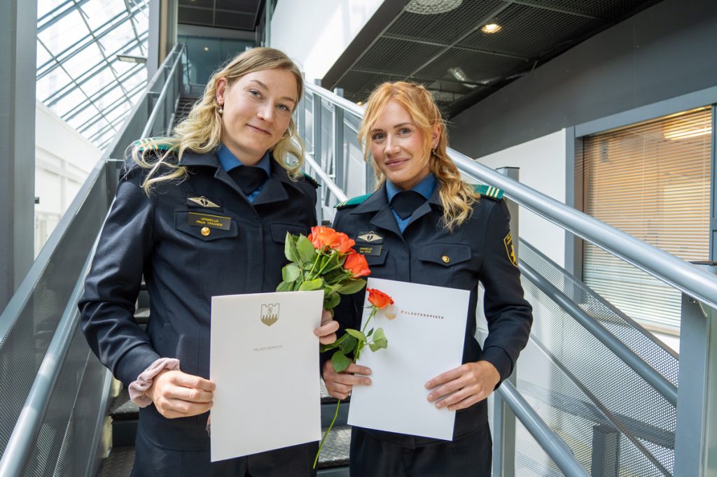 Kaksi henkilöä seisoo vierekkäin, hymyilevät ja katsovat kameraan. Käsissään heillä on tutkintotodistukset ja ruusut.
