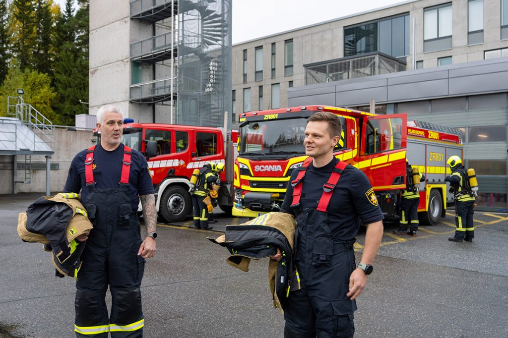 Kaksi Varsinais-Suomen hyvinvointialueen pelastuslaitoksen työntekijää Pelastusopiston kampuksella, taustalla kaksi sammutusautoa ja sammutusasuisia henkilöitä.