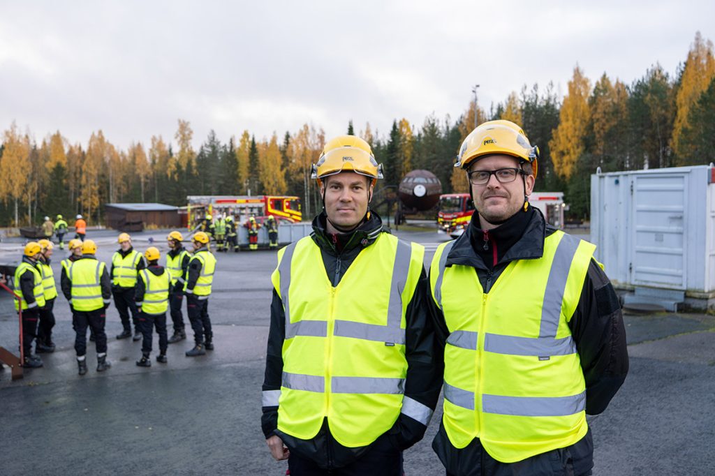 Kaksi Varsinais-Suomen hyvinvointialueen pelastuslaitoksen työntekijää Pelastusopiston harjoitusalueella, taustalla asfalttialue, suojavarusteisiin pukeutuneita henkilöitä ja sammutusautoja.